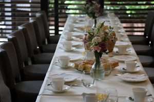 Ein elegant eingedeckter Tisch mit feinen Stofftischdecken in sanften Farben. Auf dem Tisch befindet sich ein stilvolles Blumenarrangement aus bunten Blumen, das für eine festliche Atmosphäre sorgt. Die Servietten sind kunstvoll gefaltet und harmonieren mit dem Gesamtbild. Geschirr und Besteck sind sorgfältig platziert, um eine einladende Tafel zu schaffen.