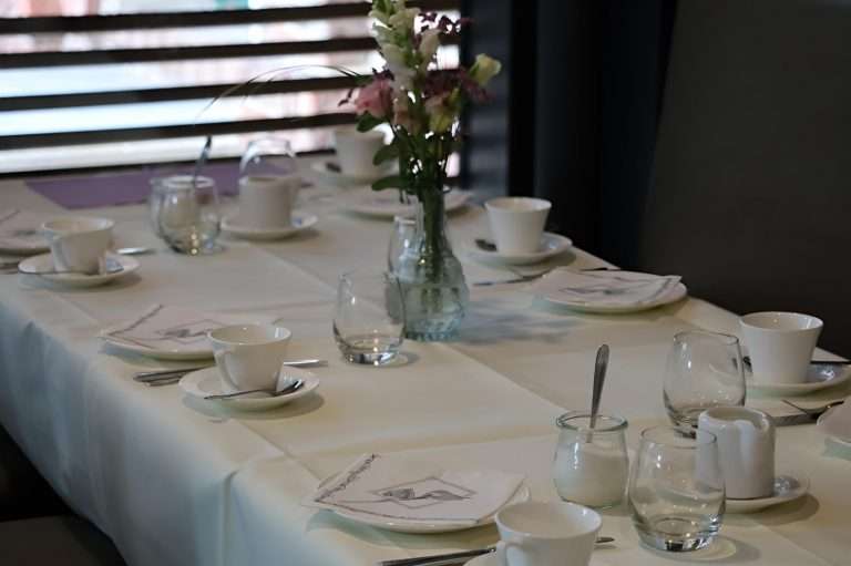 Ein elegant eingedeckter Tisch mit feinen Stofftischdecken in sanften Farben. Auf dem Tisch befindet sich ein stilvolles Blumenarrangement aus bunten Blumen, das für eine festliche Atmosphäre sorgt. Die Servietten sind kunstvoll gefaltet und harmonieren mit dem Gesamtbild. Geschirr und Besteck sind sorgfältig platziert, um eine einladende Tafel zu schaffen.